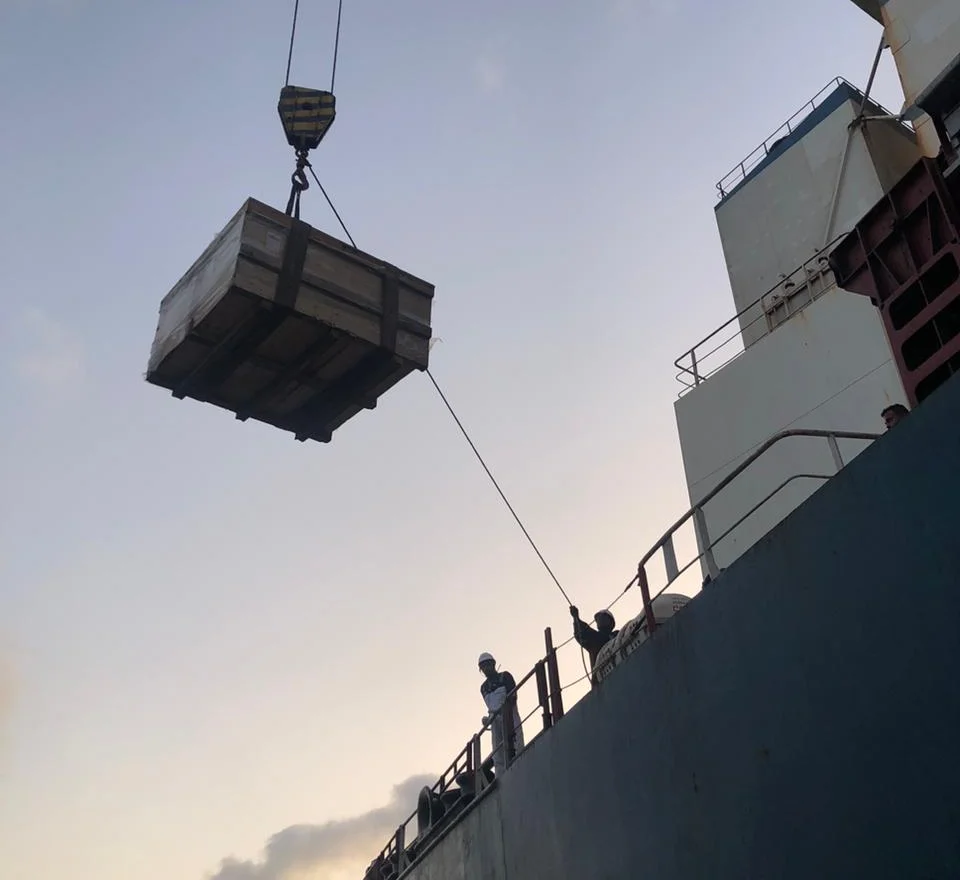 Crated equipment being craned aboard a vessel, guided by the crew on deck